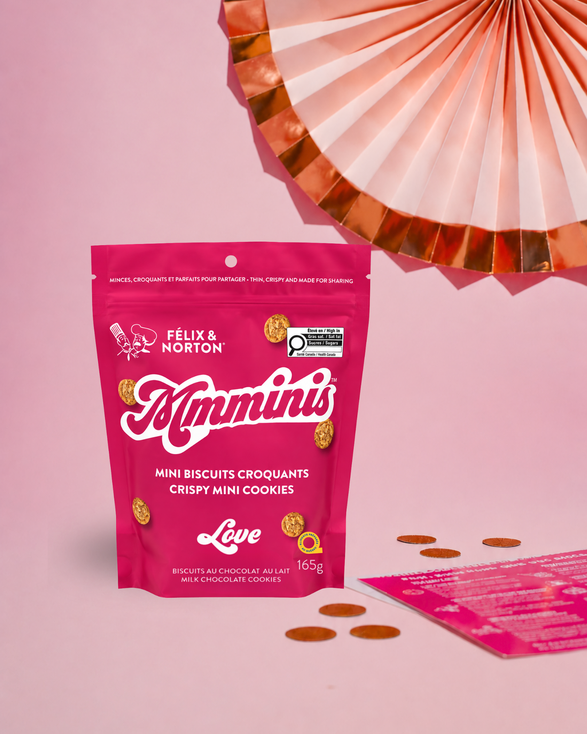 A pink stand-up pouch of Félix & Norton Minmini's Love crispy mini cookies stands on a pink surface against a pink background, with a folded paper fan and scattered mini cookies as styled props.