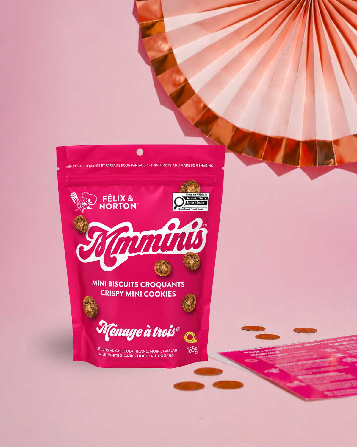A pink stand-up pouch of Felix & Norton Mimminis mini chocolate biscuits, styled on a pink background with a metallic paper fan, scattered chocolate pieces, and a branded card.